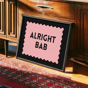 Puede incluir: Obra de arte enmarcada con el texto "ALRIGHT BAB" en letras negras sobre un fondo rosa festoneado. El marco negro se apoya en un mueble de madera sobre una alfombra estampada.