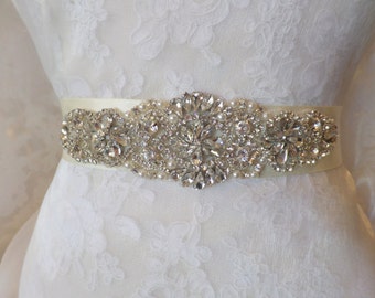 May include: A white satin bridal sash with a large, ornate rhinestone and pearl embellishment. The embellishment features a cluster of clear rhinestones and pearls in a floral design.