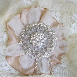 May include: A delicate, blush-coloured fabric flower with a sparkling rhinestone brooch in the centre. The flower is adorned with a sheer, white tulle overlay and is set against a white lace background.