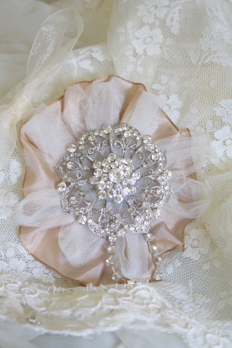 May include: A delicate, blush-coloured fabric flower with a sparkling rhinestone brooch in the centre. The flower is adorned with a sheer, white tulle overlay and is set against a white lace background.