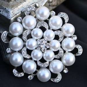 May include: A silver brooch with a floral design featuring a cluster of white pearls and clear rhinestones.