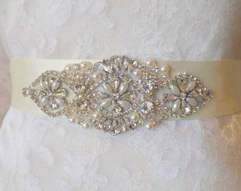 May include: A white satin bridal belt with a large, ornate rhinestone and pearl embellishment. The embellishment features a central cluster of rhinestones surrounded by smaller rhinestones and pearls.