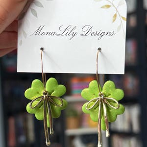 May include: A pair of green clover-shaped earrings with gold-colored bows and dangling accents. The earrings are displayed on a white card with the text "Mona Lily Designs". The background is blurred, suggesting an indoor setting.