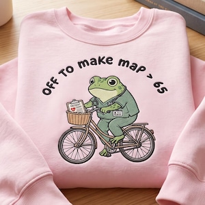 May include: A light pink crewneck sweatshirt featuring a graphic of a green frog dressed as a nurse, riding a bicycle with a basket containing a patient chart. The text "Off To make map > 65" is printed in black above the frog illustration.