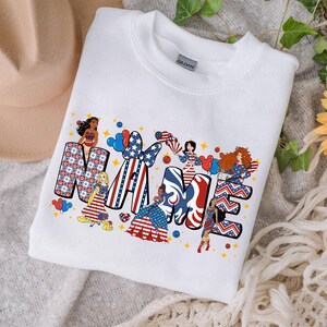 K&ouml;nnte beinhalten: Wei&szlig;es Sweatshirt mit dem Wort "NAME" in rot-wei&szlig;-blauen patriotischen Designs. Die Buchstaben sind mit Sternen, Streifen und Comicfiguren verziert. Das Sweatshirt hat einen Rundhalsausschnitt und ein Etikett mit der Aufschrift "GILDAN".