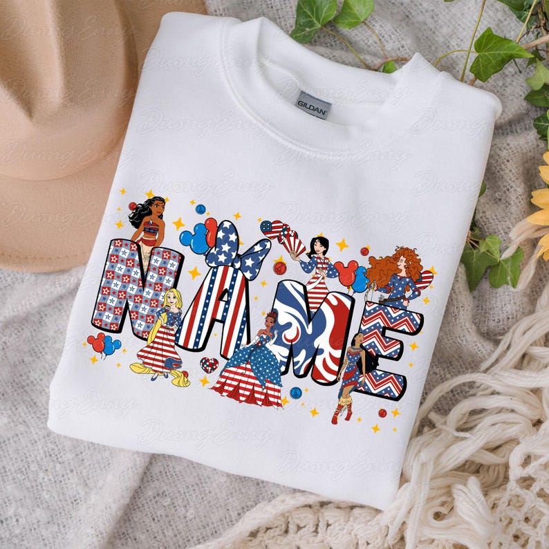 K&ouml;nnte beinhalten: Wei&szlig;es Sweatshirt mit dem Wort "NAME" in rot-wei&szlig;-blauen patriotischen Designs. Die Buchstaben sind mit Sternen, Streifen und Comicfiguren verziert. Das Sweatshirt hat einen Rundhalsausschnitt und ein Etikett mit der Aufschrift "GILDAN".
