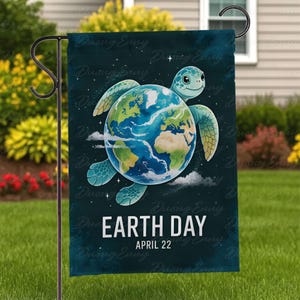 May include: A teal garden flag depicting a sea turtle with the Earth as its shell, set against a starry background. The words "Earth Day" and "April 22" are printed in white. The flag is displayed on a black metal stand in a garden.