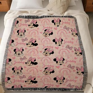 May include: Pink throw blanket with a pattern of Minnie Mouse faces, bows, rainbows, and the name "Jenny". The blanket has a fringed edge and is displayed on a bed.