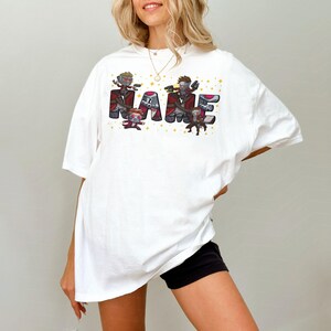 May include: White t-shirt with the word "NAME" in a colorful, cartoonish font. The letters are designed with characters holding weapons. The shirt is a casual, oversized fit.