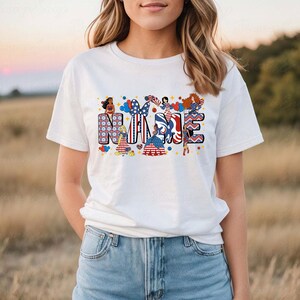 K&ouml;nnte beinhalten: Wei&szlig;es T-Shirt mit dem Wort "NAME" in einem farbenfrohen, patriotischen Design. Die Buchstaben sind mit Bildern von Frauen, Luftballons und Mustern in Rot, Wei&szlig; und Blau verziert. Das T-Shirt wird mit einer Jeans getragen.