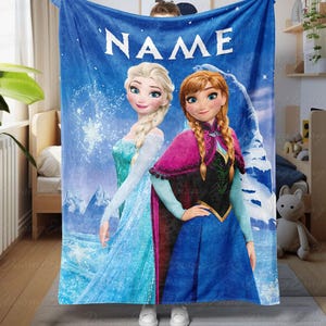 Op de afbeelding: Blauwe fleece deken met de personages Elsa en Anna uit de film Frozen. De deken heeft het woord "NAME" in witte blokletters bovenaan. De achtergrond toont een besneeuwd berglandschap.