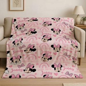 May include: A pink blanket with a repeating pattern of Minnie Mouse, bows, rainbows, and hearts. The name "Jenny" is written in a cursive font across the design. The blanket is draped over a beige sofa.