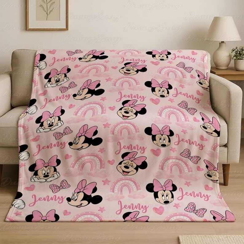 May include: A pink blanket with a repeating pattern of Minnie Mouse, bows, rainbows, and hearts. The name "Jenny" is written in a cursive font across the design. The blanket is draped over a beige sofa.
