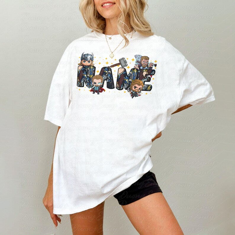 May include: White t-shirt featuring the word "NAME" in a stylized font with superhero characters. The design includes a hammer and star accents. The shirt is a casual, oversized fit.