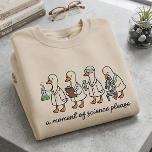 Peut inclure: Un sweat-shirt beige à col rond orné d'un motif représentant quatre canards vêtus de blouses blanches de laboratoire effectuant des tâches scientifiques, avec le texte "a moment of science please" imprimé en noir sous l'illustration.