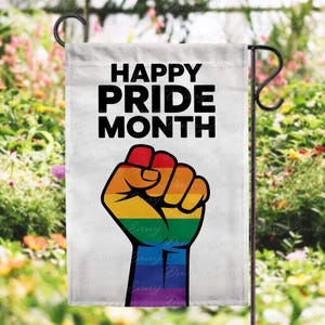 May include: White garden flag with the text "HAPPY PRIDE MONTH" in black. A rainbow-coloured fist is featured below the text. The flag is displayed on a black metal stand, with a blurred floral background.