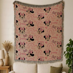 May include: A woven throw blanket with a repeating pattern of Minnie Mouse faces, bows, rainbows, and hearts in pink and black on a tan background. The name "Jenny" is written in pink script. The blanket has a fringed edge.