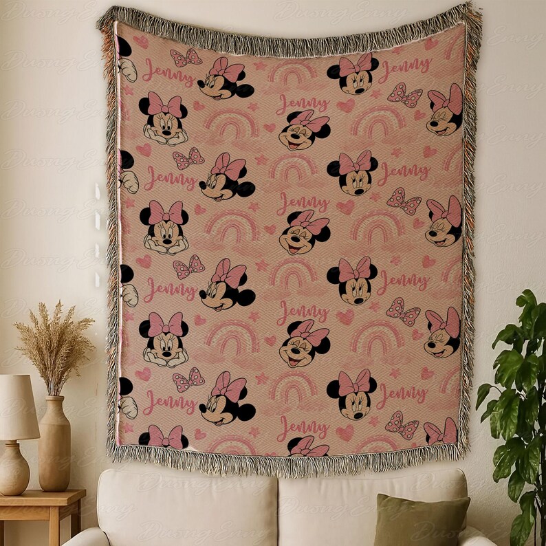 May include: A woven throw blanket with a repeating pattern of Minnie Mouse faces, bows, rainbows, and hearts in pink and black on a tan background. The name "Jenny" is written in pink script. The blanket has a fringed edge.