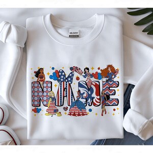 K&ouml;nnte beinhalten: Wei&szlig;es Sweatshirt mit einem patriotischen Design mit dem Wort "NAME" in Rot, Wei&szlig; und Blau. Die Buchstaben sind mit Sternen, Streifen und Cartoonfiguren verziert. Rote und blaue Luftballons sind ebenfalls enthalten.