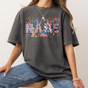 K&ouml;nnte beinhalten: Dunkelgraues T-Shirt mit dem Wort "NAME" in einem patriotischen Design. Die Buchstaben sind mit Mustern und Illustrationen von Frauen, Flaggen und Sternen in Rot, Wei&szlig; und Blau gef&uuml;llt. Das T-Shirt wird von einer Person getragen, die eine blaue Jeans tr&auml;gt.