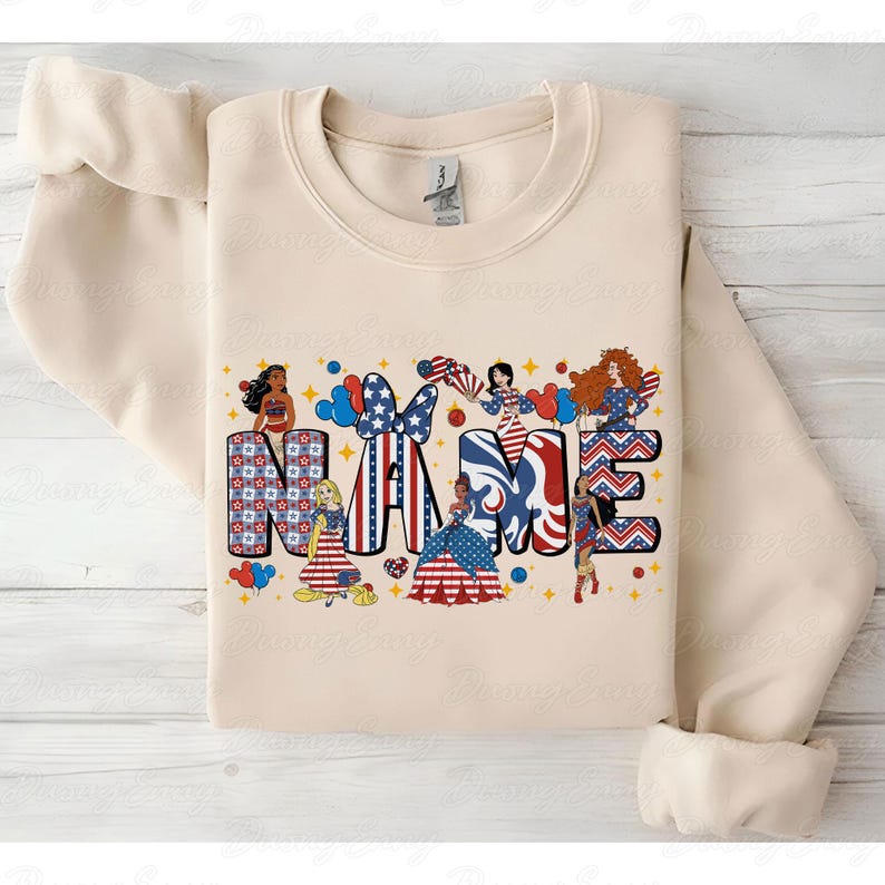 K&ouml;nnte beinhalten: Cremefarbenes Sweatshirt mit dem Wort "NAME" in einem patriotischen Design. Die Buchstaben sind mit rot-wei&szlig;-blauen Mustern und Cartoonfiguren verziert. Das Design umfasst Sterne, Streifen und Bilder von Prinzessinnen.