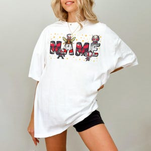 May include: White t-shirt with the word "NAME" in red and black block letters, featuring cartoon characters and gold stars. The shirt is a casual, oversized fit.