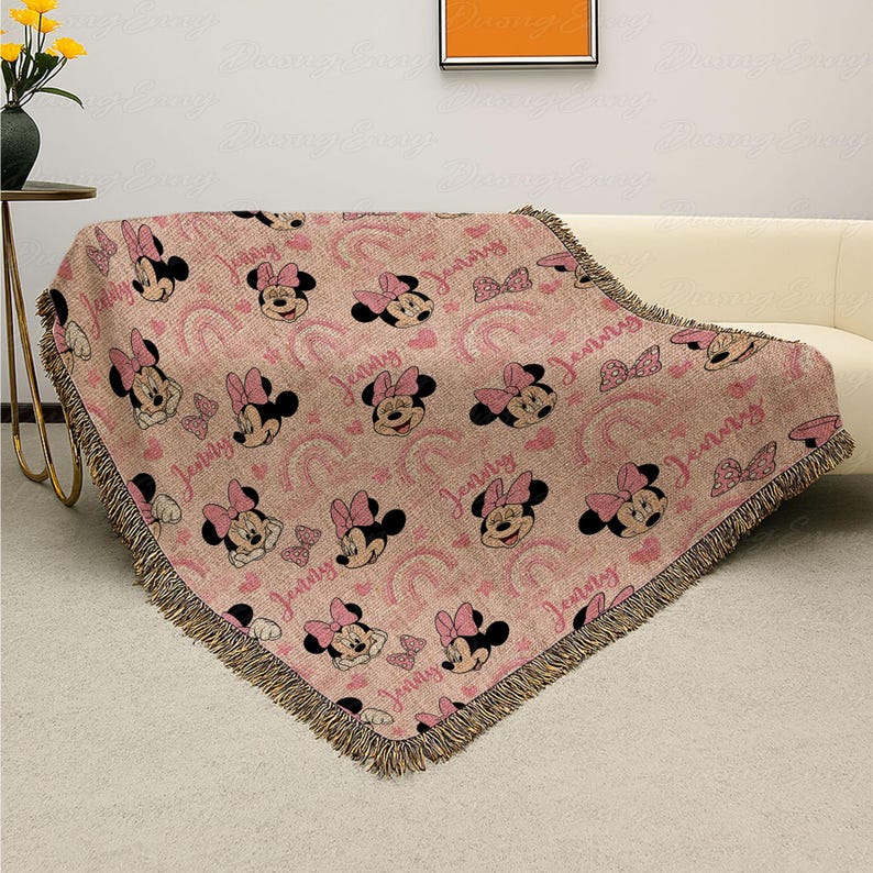 May include: Pink throw blanket with a repeating pattern of Minnie Mouse faces, bows, and rainbows. The blanket has a fringed edge and the name "Jenny" is printed throughout. It is draped over a white sofa.