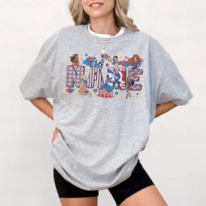 K&ouml;nnte beinhalten: Ein graues Sweatshirt mit dem Wort "NAME" in einem patriotischen Design. Die Buchstaben sind mit Illustrationen von Frauen in rot, wei&szlig; und blau gekleidet, mit Sternen und Streifen gef&uuml;llt. Das Sweatshirt hat einen Rundhalsausschnitt und lange &Auml;rmel.