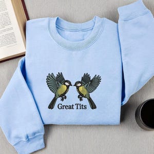 May include: A light blue long-sleeve sweatshirt featuring an embroidered graphic of two birds with yellow and grey feathers facing each other. Below the birds, the text "Great Tits" is embroidered in a dark, bold, sans-serif font.