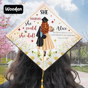Op de afbeelding: Houten afstudeerhoed met een ruitvormig ontwerp. De hoed heeft de tekst "SHE BELIEVED she could so she did!" en de naam "Alice" met universiteitsdetails. Het ontwerp bevat bloemen, sterren en een kwast.