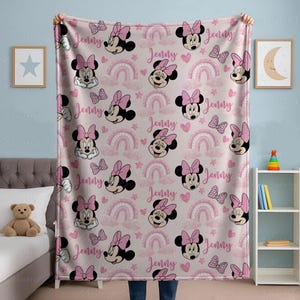 May include: Pink fleece blanket with a repeating pattern of Minnie Mouse faces, bows, rainbows, and the name "Jenny". This soft blanket is ideal for a child's bedroom or nursery, offering warmth and comfort.