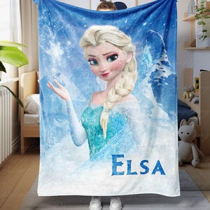 Könnte beinhalten: Eine blaue Decke mit einer Grafik von Elsa aus dem Film Frozen. Elsa wird mit blonden Haaren und einem blauen Kleid vor einem schneebedeckten Hintergrund dargestellt. Der Name "ELSA" ist unten in Blau gedruckt.