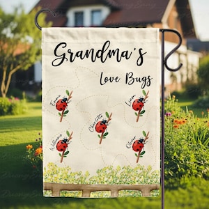 May include: A beige garden flag with the words "Grandma's Love Bugs" in black script. It features ladybug illustrations with names like Emma, Liam, and Charlotte. A wooden fence and greenery are at the bottom.