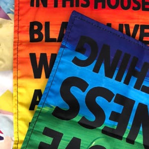 May include: Close-up of three colorful flags with bold black text. The flags display phrases like "Black Lives Matter" and "Love Wins" against vibrant gradients of orange, blue, and green. The flags are likely for outdoor display.