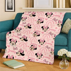 May include: A pink throw blanket with a repeating pattern of Minnie Mouse, bows, rainbows, butterflies, and hearts. The name "Jenny" is written in pink script. The blanket is draped over a teal sofa.