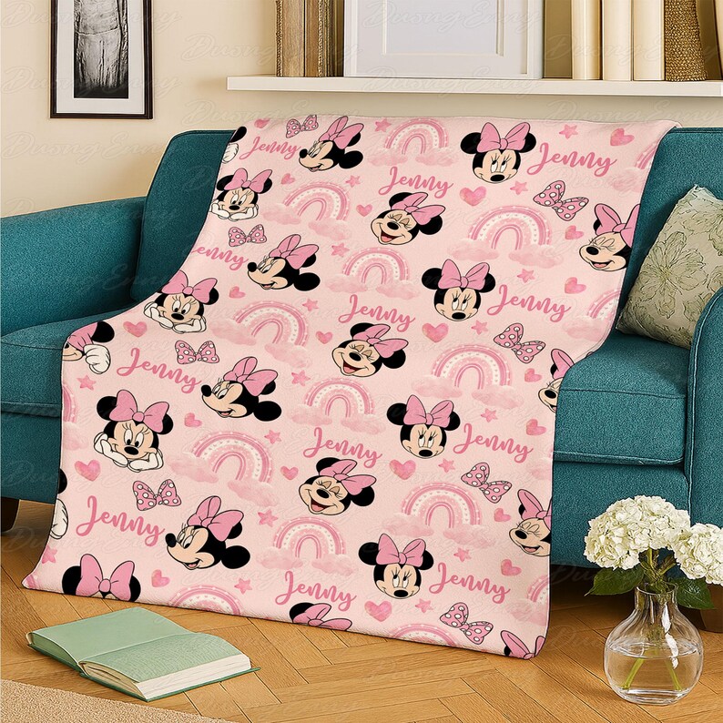 May include: A pink throw blanket with a repeating pattern of Minnie Mouse, bows, rainbows, butterflies, and hearts. The name "Jenny" is written in pink script. The blanket is draped over a teal sofa.