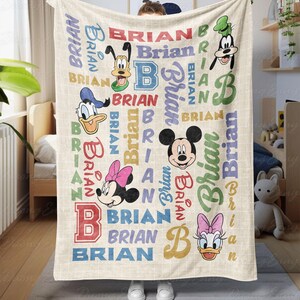 Könnte beinhalten: Eine beige Decke mit dem Namen "Brian" in verschiedenen bunten Schriftarten und Disney-Charakterillustrationen. Die Decke enthält Bilder von Mickey Mouse, Minnie Mouse, Donald Duck, Daisy Duck und Goofy. Die Decke wird von einer Person gehalten.