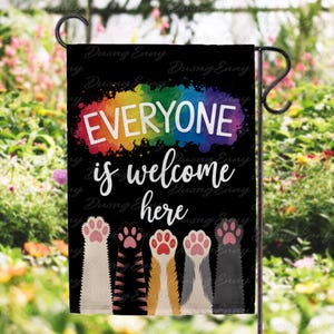 Peut inclure: Drapeau de jardin noir avec le texte "EVERYONE is welcome here" en blanc. Le texte est au-dessus d'une rangée de pattes de chat colorées. Une éclaboussure de peinture aux couleurs de l'arc-en-ciel est derrière le mot "EVERYONE".