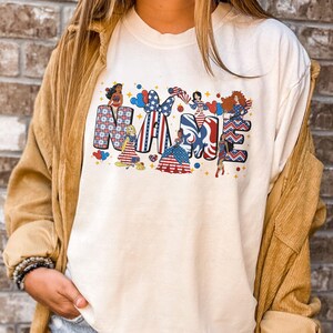 K&ouml;nnte beinhalten: Cremefarbenes T-Shirt mit dem Wort "MAMA" in einem patriotischen Design. Jeder Buchstabe ist mit Mustern und Illustrationen von Frauen in rot, wei&szlig; und blauen Kleidern mit Sternen und Streifen verziert.
