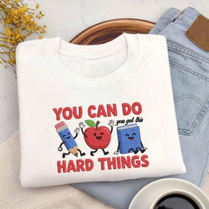 May include: A white crewneck sweatshirt featuring an embroidered design of a blue pencil, a red apple, and a blue book with smiling faces. The text reads YOU CAN DO you got this HARD THINGS in red and black thread, centred on the chest.