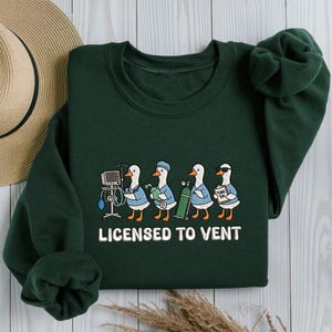 May include: A dark green long-sleeve sweatshirt featuring a graphic of four white ducks dressed in blue medical scrubs. The ducks are depicted with medical equipment including a monitor, oxygen tank, and clipboard. The text reads LICENSED TO VENT.