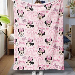 May include: Pink blanket with a pattern of Minnie Mouse, bows, rainbows, hearts, and stars. The name "Jenny" is printed across the design. A soft, cosy blanket suitable for children's bedrooms or nurseries.