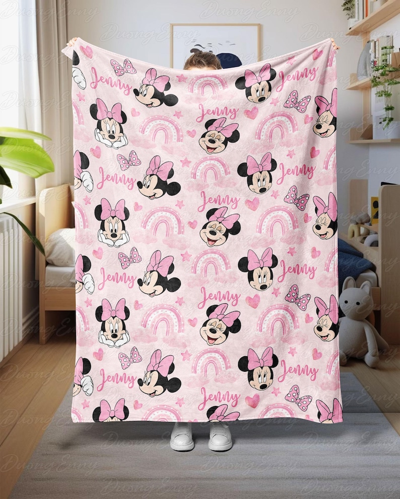 May include: Pink blanket with a pattern of Minnie Mouse, bows, rainbows, hearts, and stars. The name "Jenny" is printed across the design. A soft, cosy blanket suitable for children's bedrooms or nurseries.