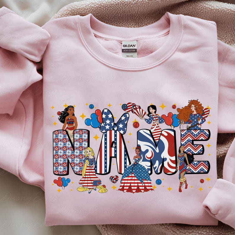 K&ouml;nnte beinhalten: Ein hellrosa Sweatshirt mit dem Wort "NAME" in einem patriotischen Design. Die Buchstaben sind mit Sternen und Streifen verziert und von Zeichentrickfiguren umgeben. Das Sweatshirt ist aus einem weichen Material.