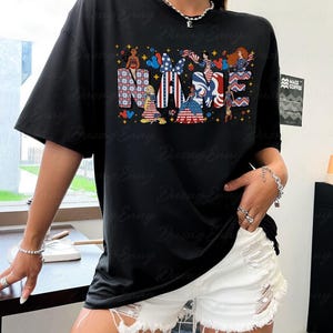 K&ouml;nnte beinhalten: Schwarzes T-Shirt mit dem Wort "NAME" in einem patriotischen Design mit Cartoon-Charakteren und Elementen der amerikanischen Flagge. Das Shirt hat kurze &Auml;rmel und eine lockere Passform.