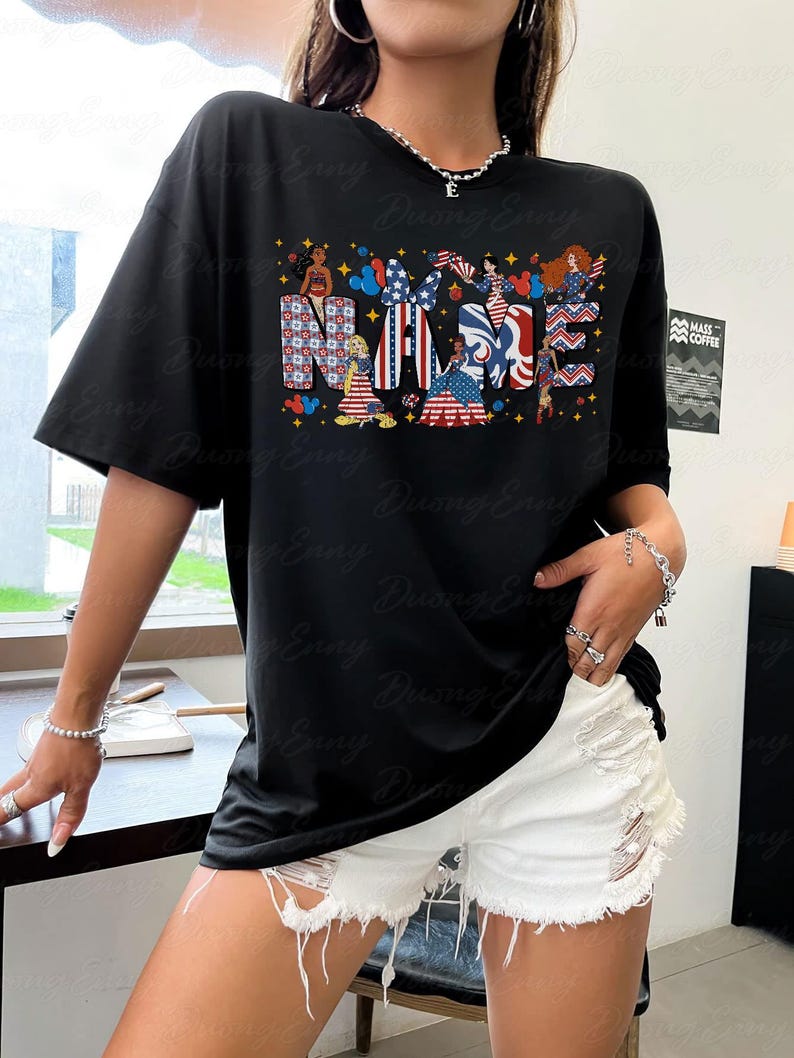 K&ouml;nnte beinhalten: Schwarzes T-Shirt mit dem Wort "NAME" in einem patriotischen Design mit Cartoon-Charakteren und Elementen der amerikanischen Flagge. Das Shirt hat kurze &Auml;rmel und eine lockere Passform.