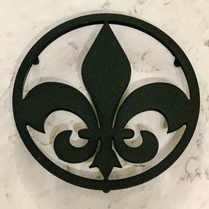 Könnte beinhalten: Schwarzes Metall-Fleur-de-Lis-Design in einem kreisförmigen Rahmen. Das dekorative Stück zeigt ein stilisiertes Lilien-Symbol, ein klassisches Emblem. Das Design ist vor einem weiß-grauen marmorähnlichen Hintergrund platziert.
