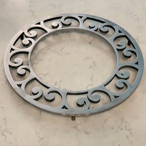 Puede incluir: Un salvamanteles decorativo circular de metal con un intrincado diseño calado. El salvamanteles presenta una serie de patrones arremolinados en forma de volutas alrededor del borde exterior y una abertura central. El metal tiene un acabado gris plateado.