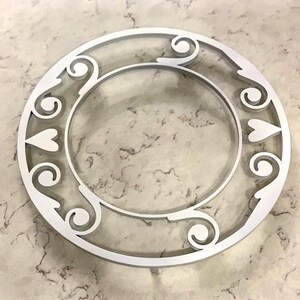 May include: A white, circular decorative object with an open center. The design features heart shapes and swirling patterns around the outer edge. The object is made of a solid material and sits on a light-colored, textured surface.