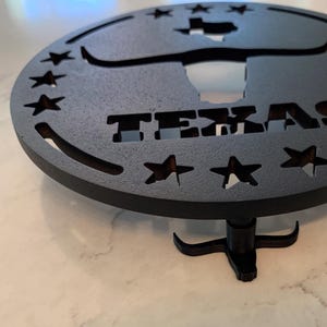 Peut inclure: Pièce décorative en métal noir en forme de Texas, avec un design circulaire. Le mot "TEXAS" est découpé dans le métal, ainsi qu'une silhouette de l'État et des étoiles. Il est soutenu par un petit support noir.